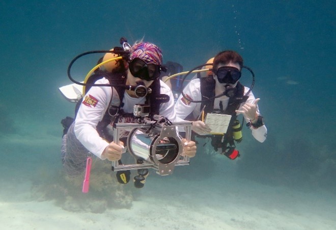 Two divers in the water wearing white shirts with the REEF logo on the upper arm. One holds a large underwater camera, the other holds a pad and pencil while showing the ‘hang loose’ hand signal.