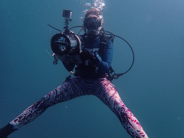 A female diver in red patterned pants has her legs wide while holding a large underwater camera. 